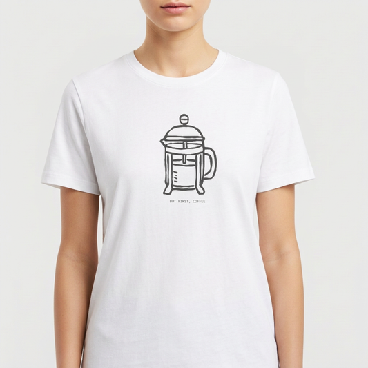 Camiseta com estampa frontal de cafeteira tipo prensa francesa desenhada de forma minimalista, acompanhada da frase "BUT FIRST, COFFEE" em letras discretas.